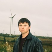 A guy in a brown jumper and black leather jacket stood in a field in front of wind turbines